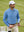 A male model wearing a light blue long sleeve herringbone polo shirt, white golf cap, and cream trousers while holding a tennis racket on a green outdoor court.